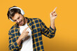 Portrait of handsome beard guy in white t-shirt and plaid shirt listening to music in headphones singing and pointing fingers aside isolated on yellow orange background