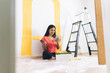 © Westend61 - Smiling young woman with paint roller using smart phone in living room