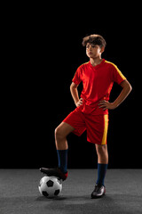 Wall Mural - Full-length portrait of young boy, teenager, professional football player in uniform with ball posing isolated over black background