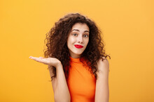 Well Sorry. Unaware And Clueless Silly Charming Woman With Curly Hairstyle Shrugging And Smirking With Uncertain Look Raising Palm In Who Knows Gesture, Posing Uninvolved Over Orange Background