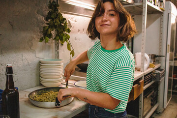 happy young woman cooking spaghetti with pesto hands holding pasta bunch putting into the frying pan to mix with pesto sauce and white beans vegan recipe preparing food smiling at camera