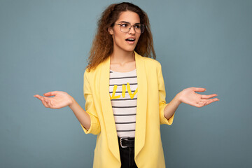 Wall Mural - Photo of young dissatisfied astonished shocked beautiful brunette curly woman with sincere emotions wearing casual yellow jacket and optical glasses isolated on blue background with copy space and