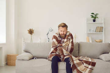 It's cold at home in wintertime. Man freezing in his house in winter because of broken thermostat. Sad bearded young guy wrapped in woolen plaid shivering while sitting on sofa in living room interior