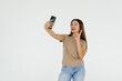 Attractive young woman holding mobile phone and making photo of herself while standing against white background