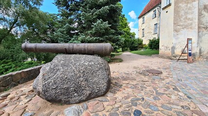 An old artillery cannon is installed on large gray stone at the gate of Latvian Jaunpils Castle in the summer of 2021