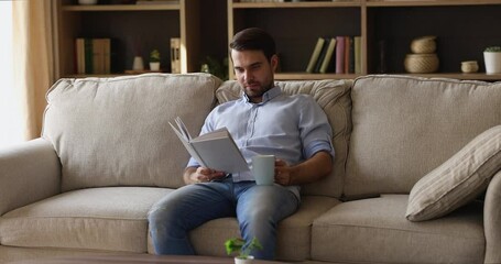 Wall Mural - Best way to spend leisure time. Peaceful young male introvert pass free serene weekend on cozy sofa at home read favorite book drink hot tea. Calm carefree man enjoy favorite novel with cup of coffee