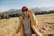 Young blond woman with sunglasses dancing in mountains. Freedom, happiness, travel and vacations concept, outdoor activities. Happy Girl wearing red hat and a yellow jacket