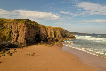 Poster - Beautiful view of a beach with high green hills next to wavy water under a blue sunny cloudy sky