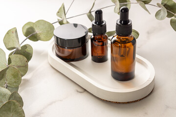 anti-aging collagen facial serum in dark glass bottle and face cream on white concrete tray on marble background with eucalyptus branch. Natural Organic Cosmetic Beauty Concept.