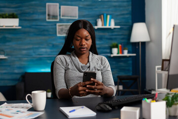 Poster - African american woman using mobile phone to work from home. Young entrepreneur holding smartphone and looking at screen, working on remote business project at desk with computer.