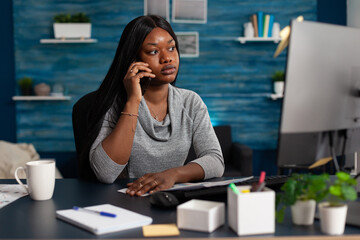 Poster - African american woman talking on phone doing remote work on computer monitor at home. Entrepreneur talking about business project on smartphone, designing data charts on software