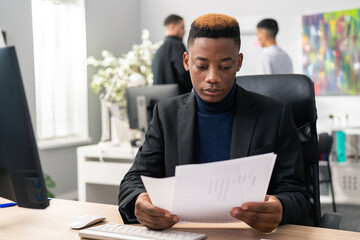 Wall Mural - A young boy with unusual hair sits on a chair at a desk in a company, in his hands he holds reports, documents, reads carefully, analyzes the notes, focused, attentive, serious employee in a jacket