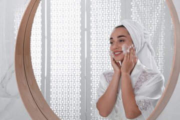 Poster - Beautiful young woman applying cleansing foam onto face near mirror in bathroom. Skin care cosmetic