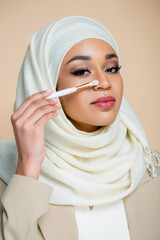 Wall Mural - young muslim woman in hijab holding cosmetic brush near face isolated on beige