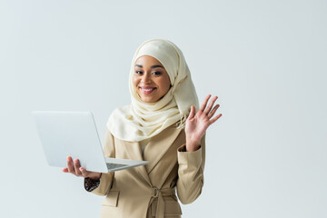 cheerful muslim woman in hijab and beige suit holding laptop and waving hand isolated on grey