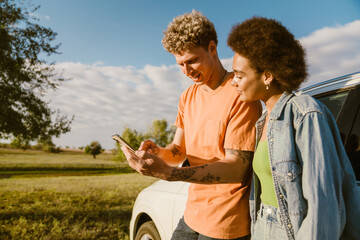 Young multiracial couple using cellphone and standing by car during trip