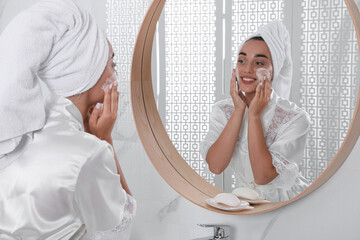 Poster - Beautiful young woman applying cleansing foam onto face near mirror in bathroom. Skin care cosmetic