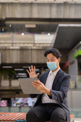 Wall Mural - Asian businessman wearing face mask and sitting outdoors in city using digital tablet computer