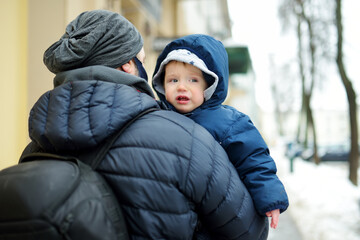 Wall Mural - Cute little baby boy in his fathers arms. Dad and son having fun on chilly winter day in city park.