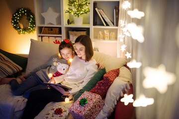 Wall Mural - Two cute young sisters using a tablet pc at home in warm and cozy living room at Christmas. Family having online video call on Xmas eve.