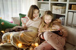 © MNStudio - Two young sisters snuggling up on the sofa in a cozy living room at Christmas. Cute children using a tablet at home during winter break. Kids reading a book in comfy blanket.