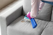 Woman in gloves cleaning grey sofa with brush and detergent at home