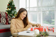 © chinnarach - close up young asian woman try to put ribbon on wrapped gift box at living room with pine tree decoration for merry Christmas and happy new year festival season concept