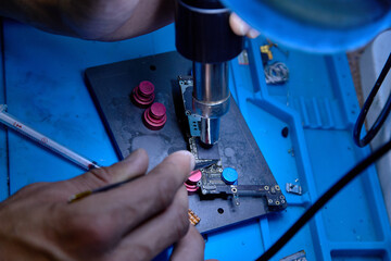  Hands of professional technician repairman work on chipset pins of removed microprocessor from smartphone motherboard with tweezers and hot air heater. Close-up macro photo, Selective focus