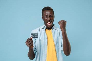 Wall Mural - Screaming young african american man guy in casual shirt, yellow t-shirt posing isolated on blue background. People lifestyle concept. Mock up copy space. Hold credit bank card doing winner gesture.