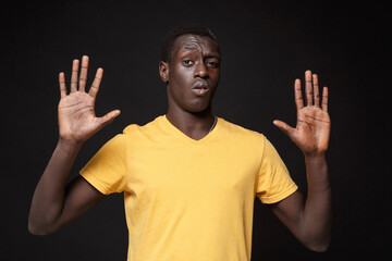 Wall Mural - Perplexed young african american man guy in yellow t-shirt posing isolated on black background studio portrait. People emotions lifestyle concept. Mock up copy space. Rising hands up, showing palms.