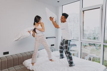 Wall Mural - Full size body length excited young couple two friends man woman family in casual white clothes stand on bed do pillow fight relax spend time together in bedroom lounge home house wake up good day