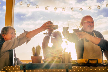 Wall Mural - Old senior group of caucasian people enjoy nd celebrate together clinking with red wine during bbq grill at home in terrace. Sun sunlight in background. Cheerful men and women elderly lifestyle