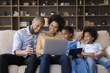 Happy young african american couple parents and cute little children using different gadgets, spending time online, playing games, entertaining sitting together on sofa in living room, tech addiction.