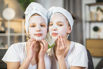 Portrait of pretty female teenagers posing at living room with hydration mask on face. Two sisters taking care of young skin using various cosmetics.