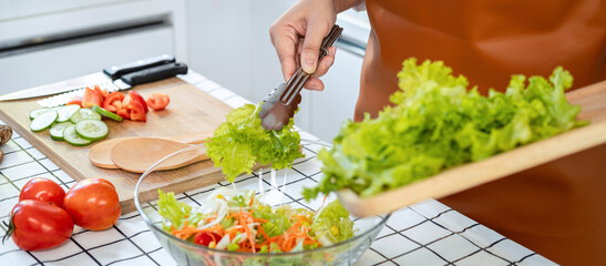 Wall Mural - Close up of asian housewife wearing apron and holding chopping board with lettuce to mixing with cucumber