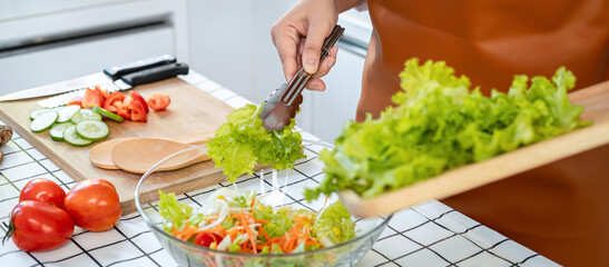 Poster - Close up of asian housewife wearing apron and holding chopping board with lettuce to mixing with cucumber