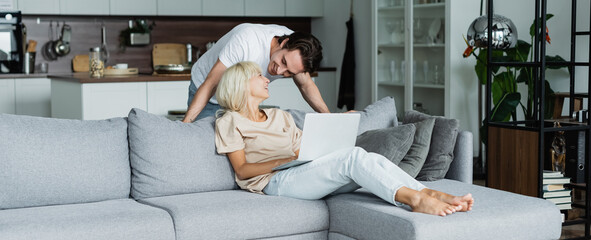 happy man looking at blonde girlfriend working from home with laptop, banner