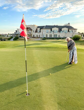 Senior Woman Plays Golf On Golf Course. Background Of Luxury Mansion. Scores Ball In Hole With Flag
