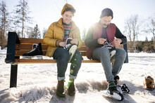 Gay Male Couple Putting On Ice Skates On Bench In Snowy Winter Park