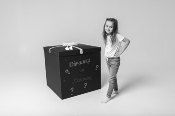 Canvas Print - Black and white photo of a little cute emotional girl in a colored dress with blond hair leaned on a festive black box with a bow on a white background. She is thoughtful, smiling in anticipation.