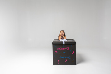 Canvas Print - little cute emotional girl with blond hair leaned on a festive black box with a bow on a white background. She is thoughtful, smiling. Birthday, celebration, anticipation