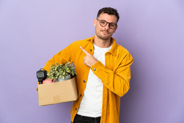 Poster - Young caucasian man making a move while picking up a box full of things isolated on purple background pointing to the side to present a product