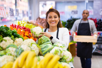 Latin saleswoman in black apron and her assistant with box of vegetables in background