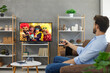 © Studio Romantic - Man watching movie or entertaining TV show at home. Young guy sitting on couch and switching on television set with big HD screen that is standing on wall rack in modern living room interior