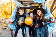 © klavdiyav - siblings boy in skeleton costume, teenage girl in witch costume and hat and cute little girl in spooky costume sits in trunk car decorated for Halloween with web, orange balloons and pumpkins, outdoor