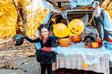 Little Girl In Spooky Cute Costume And Hat Stands Near Trunk Car Decorated For Halloween With Web, Orange Balloons And Pumpkins, Outdoor Creative Activity Concept In Autumn