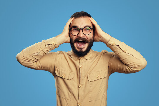 Shocked bearded man screaming while holding hands on head, looking with bugged eyes and opened mouth, realizing he won lottery, isolated over blue 