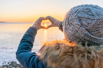 Poster - A girl in winter clothes makes a heart with her hands in gloves. Against the background of the frozen sea at sunset