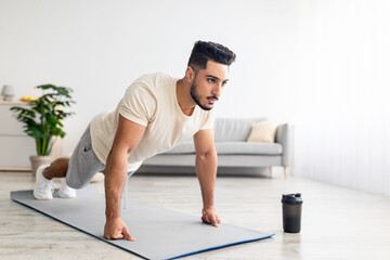 Wall Mural - Domestic sports. Athletic young Arab man standing in plank pose, doing push ups, working out core muscles at home