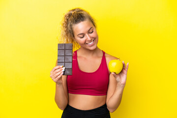 Girl with curly hair isolated on yellow background taking a chocolate tablet in one hand and an apple in the other